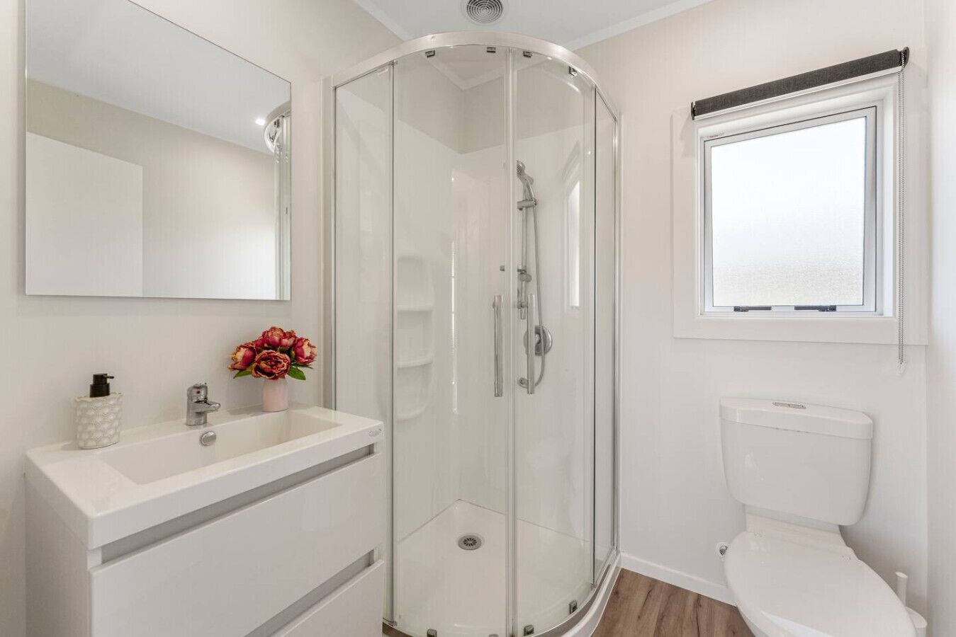 A modern bathroom with a glass-enclosed shower, a white sink with a vase of flowers, and a toilet with a window above it.