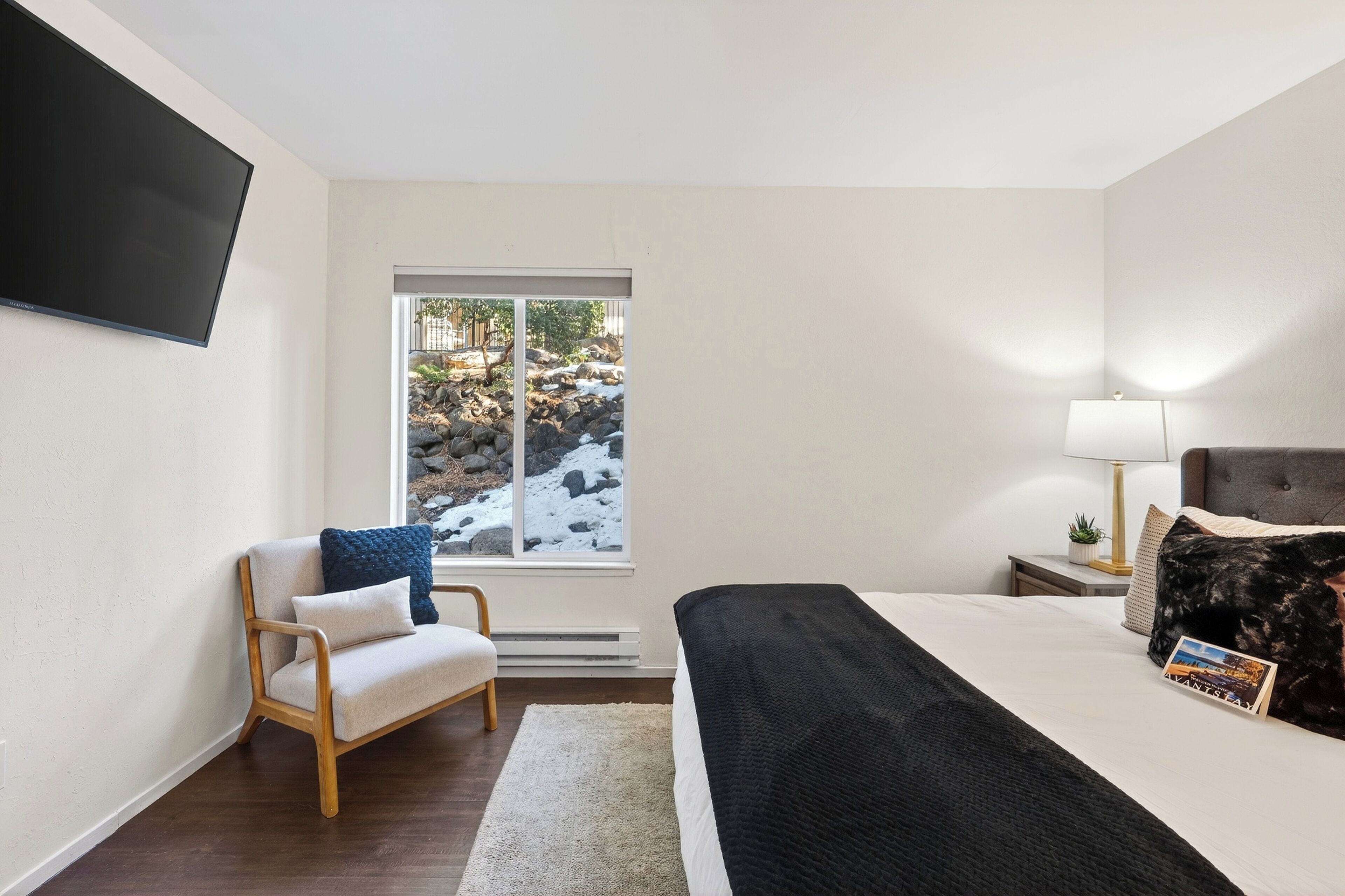 Ein modernes Hotelzimmer mit einem großen Bett, einem Sessel, einem Fernseher, einem Fenster mit Blick auf Felsen und Grünflächen und einem Nachttisch mit Lampe.