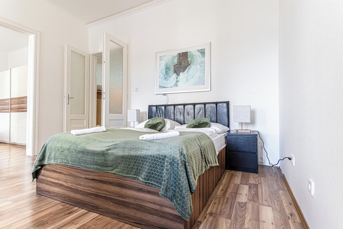 A bedroom with a wooden bed, green bedding, a nightstand, a lamp, and a painting on the wall.