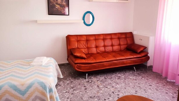 Ein Zimmer mit einem roten Sofa, einem Bett mit einem gemusterten Überwurf, einem gerahmten Bild an der Wand und einem kleinen Topfpflanzen auf einem Regal.