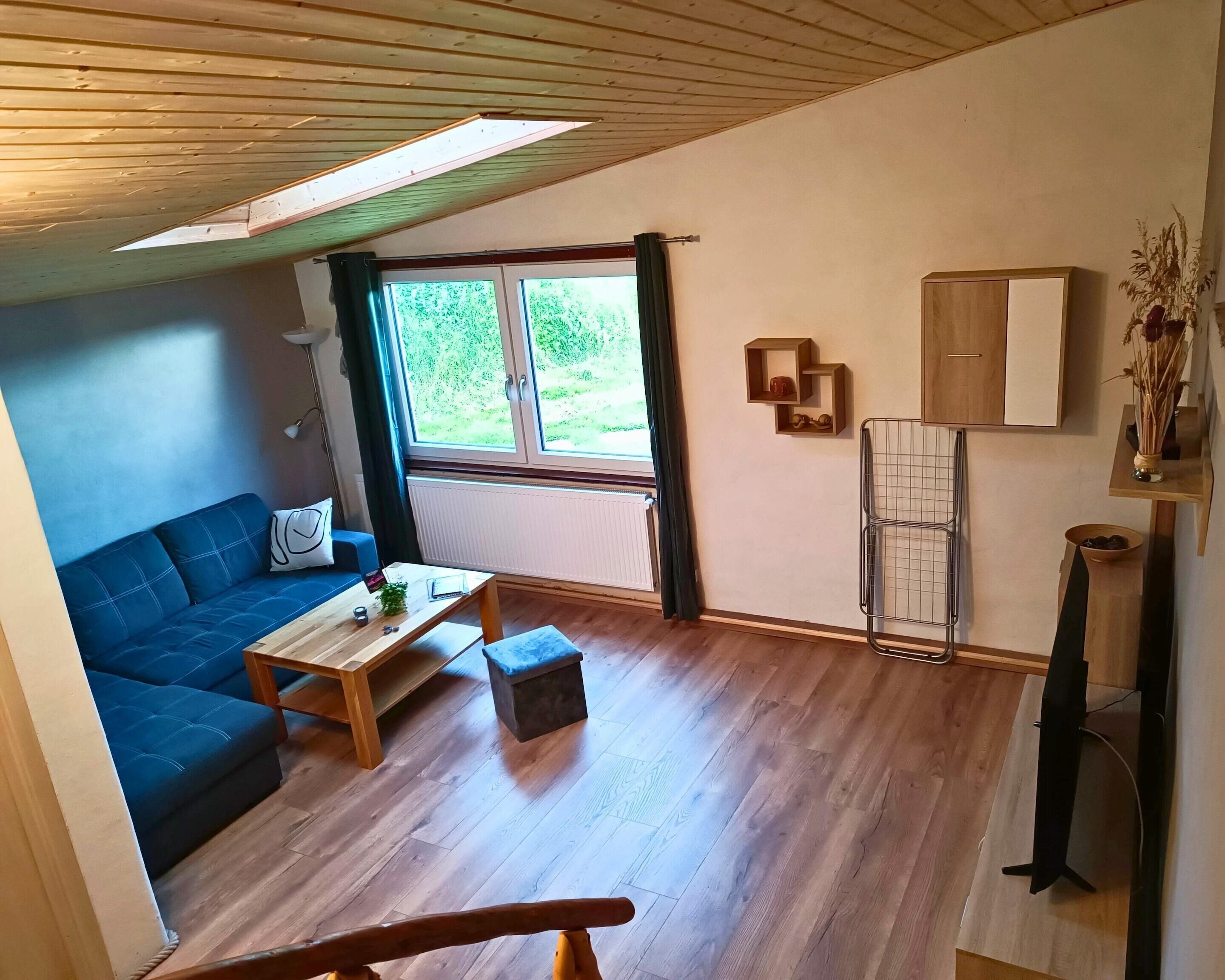 A cozy living room with a blue sofa, wooden coffee table, and a small table with a plant. A window with curtains offers a view of greenery outside.