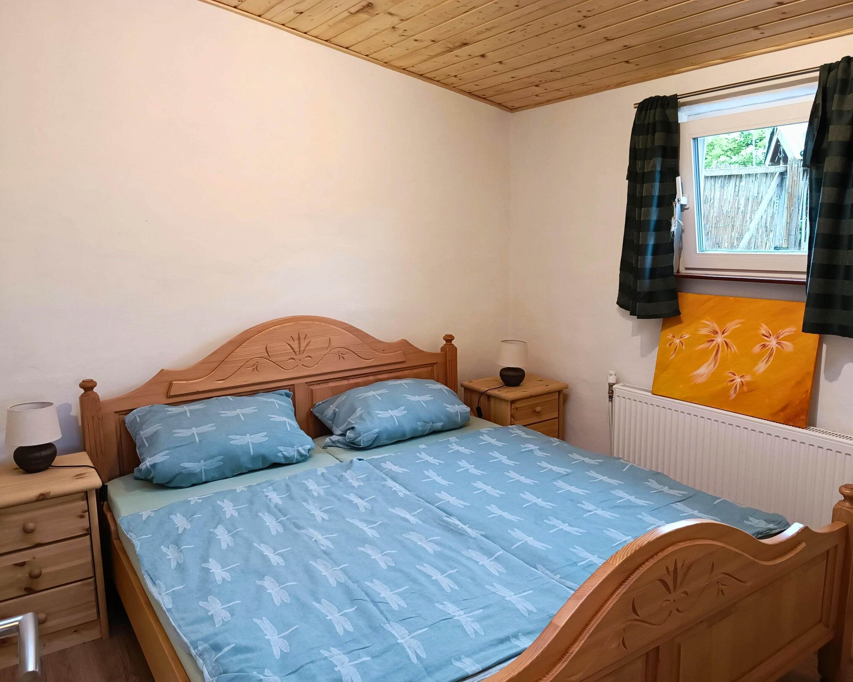 Un dormitorio con cama de madera, dos almohadas, una mesita de noche con lámpara, una ventana con cortinas y un radiador.