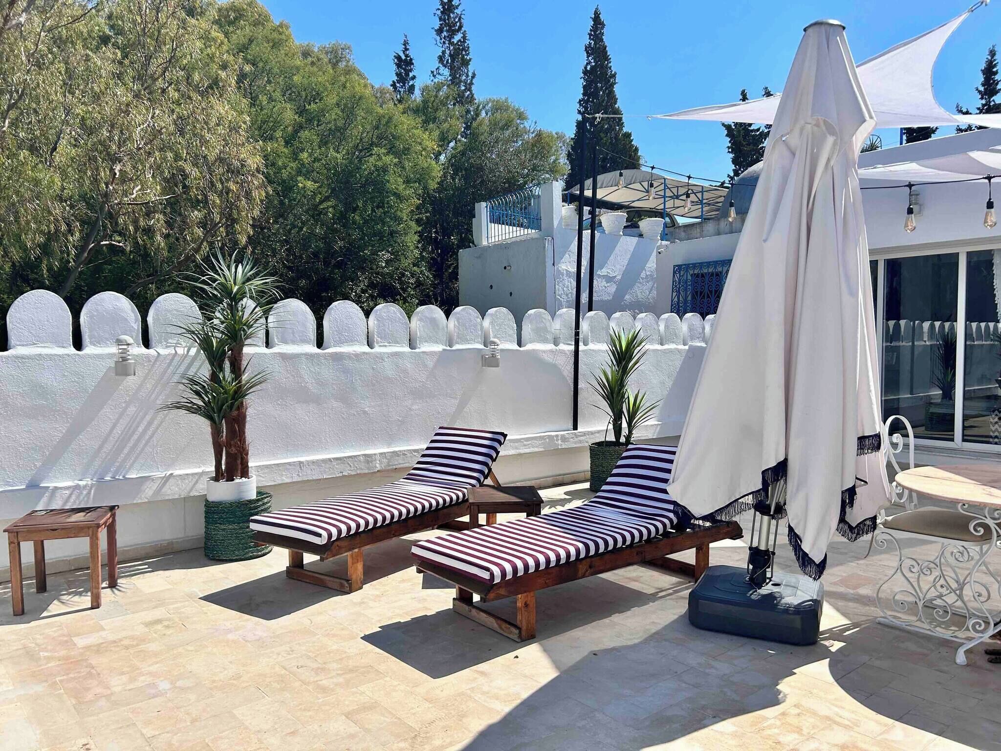 Un espace extérieur ensoleillé, aménagé avec deux fauteuils et une table, entouré de murs blancs et d’une clôture blanche.