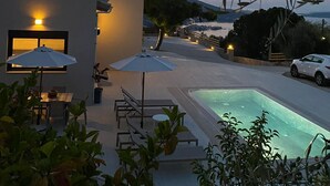 Una casa moderna con piscina, mobiliario exterior y vista al mar al anochecer.