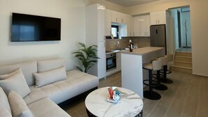 Una sala moderna con un sofá, una mesa de centro redonda y un televisor. Hay una zona de cocina con una barra y taburetes.
