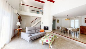 Un amplio salón con un sofá gris, una mesita pequeña y un camino de mesa floral. Una escalera conecta con un nivel superior.