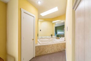 Ein Badezimmer mit einer großen Badewanne, einer Tür und einem Spiegel, der ein Bett und eine Pflanze reflektiert.