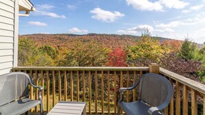 Eine Holzterrasse mit zwei Metallstühlen und einem Tisch, mit Blick auf einen bewaldeten Hügel im Herbstlaub.