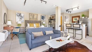 Ein modernes Wohnzimmer mit einer Couch, einem Couchtisch und einer kleinen Küchenzeile inklusive Kühlschrank und Barhocker.