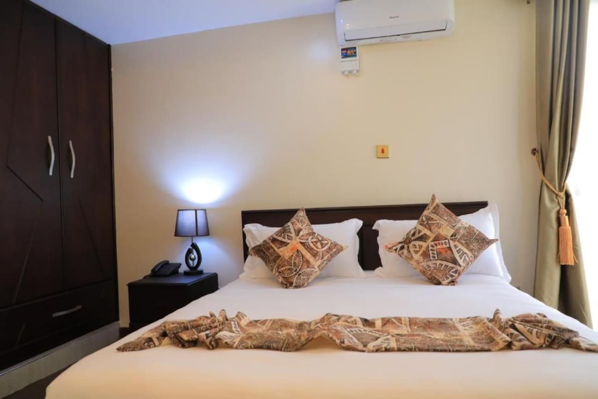 A hotel room with a bed, a nightstand, a lamp, an air conditioning unit, and a wardrobe.