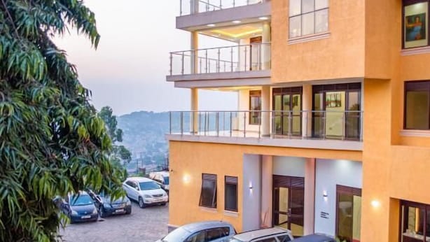 A multi-story building with a yellow facade, multiple balconies, and a parking area with several cars. - Precious villas hotel  (Kampala)