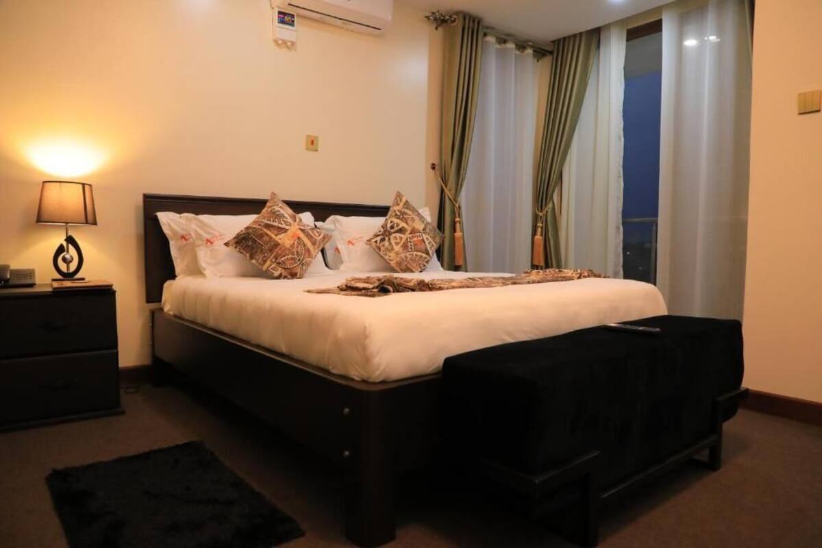A hotel room with a bed, a bedside table with a lamp, a black ottoman, and a view of the outside through curtains.