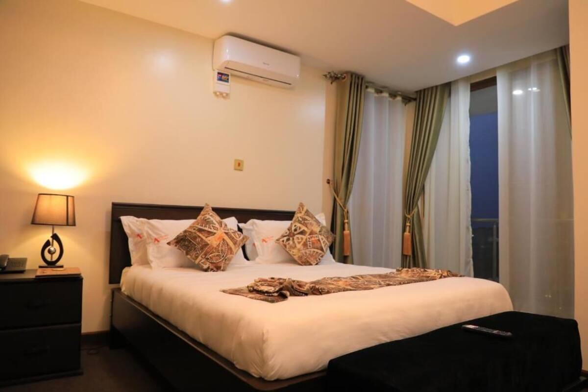 A hotel room with a bed, a nightstand, a lamp, an air conditioner, and a window with curtains.