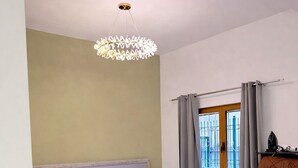 Habitación de hotel con cama, televisor, mesa de comedor con sillas, araña y ventana con cortinas.