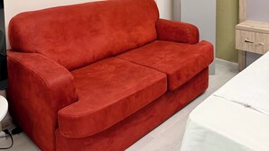 Una habitación con un sofá rojo, una cama blanca, una mesita de madera y un armario gris.