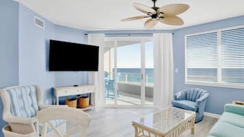 A living room with a flat-screen TV, wicker furniture, a ceiling fan, and a view of the ocean through large windows.