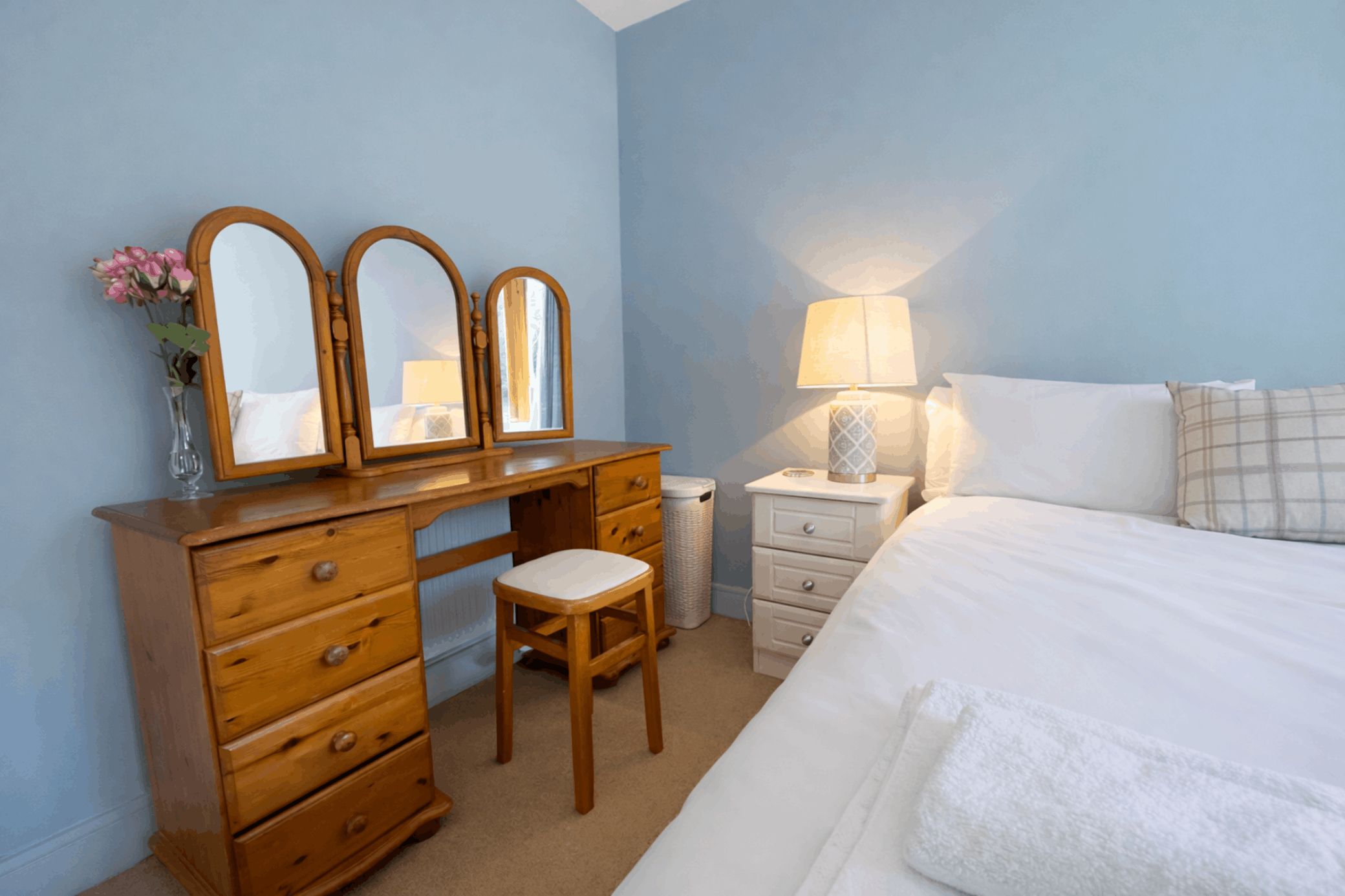 A bedroom with a wooden dresser, a chair, a bedside table with a lamp, and a bed with white bedding.