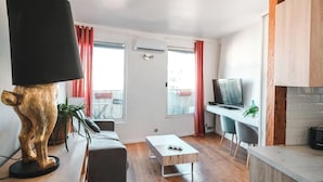 Una sala de estar moderna con sofá, mesa de centro y televisor sobre un mueble de madera. Se aprecia un amplio ventanal con cortinas y un balcón.