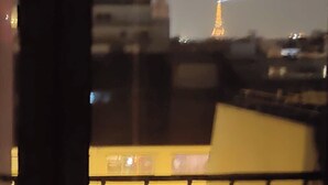 Vista a través de una ventana con barandilla, que muestra un edificio con un ventanal iluminado y una torre lejana iluminada.