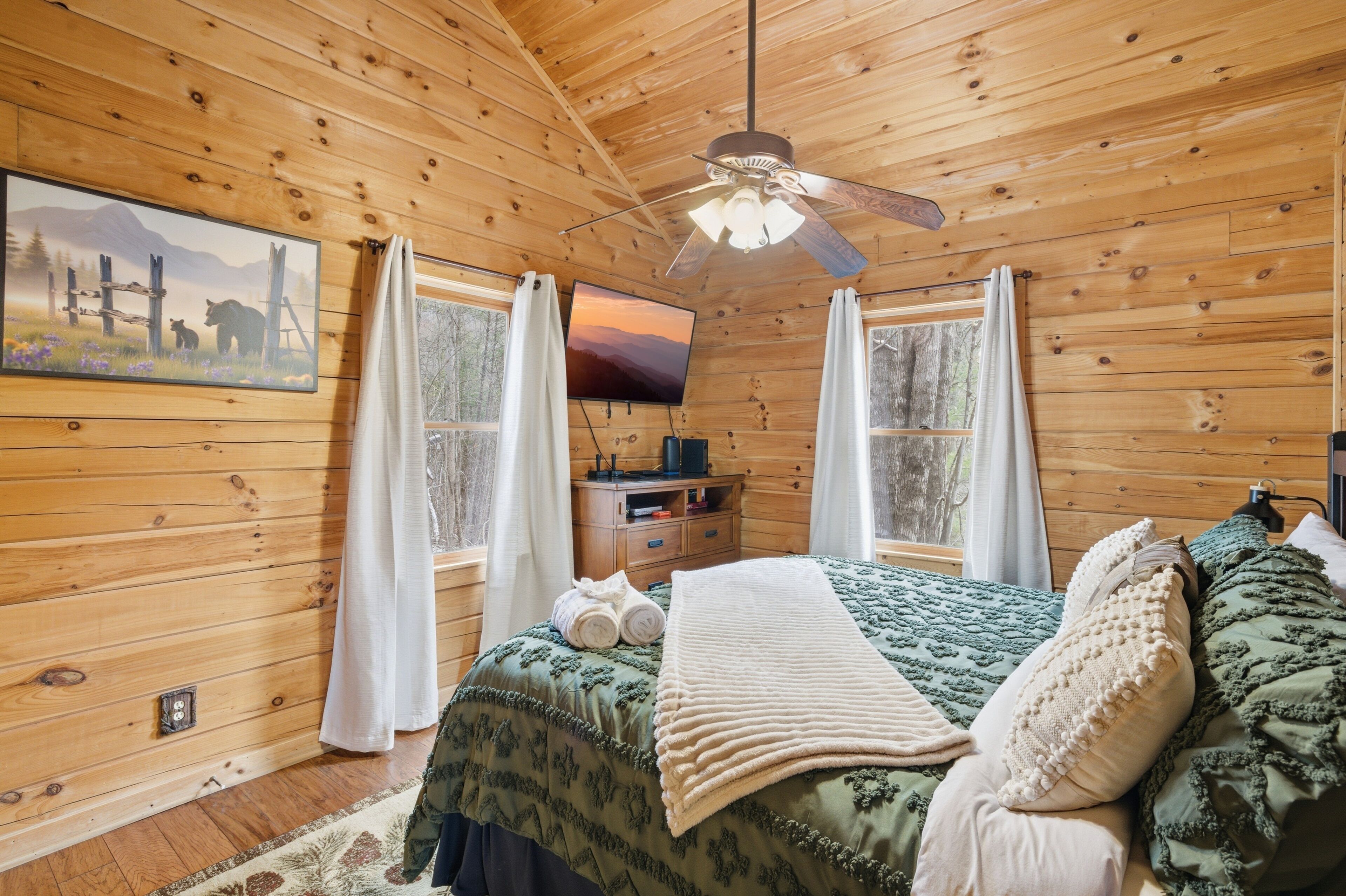 A wooden cabin bedroom with a bed, a ceiling fan, a TV, and a painting on the wall.