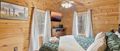 A wooden cabin bedroom with a bed, a ceiling fan, a TV, and a painting on the wall.