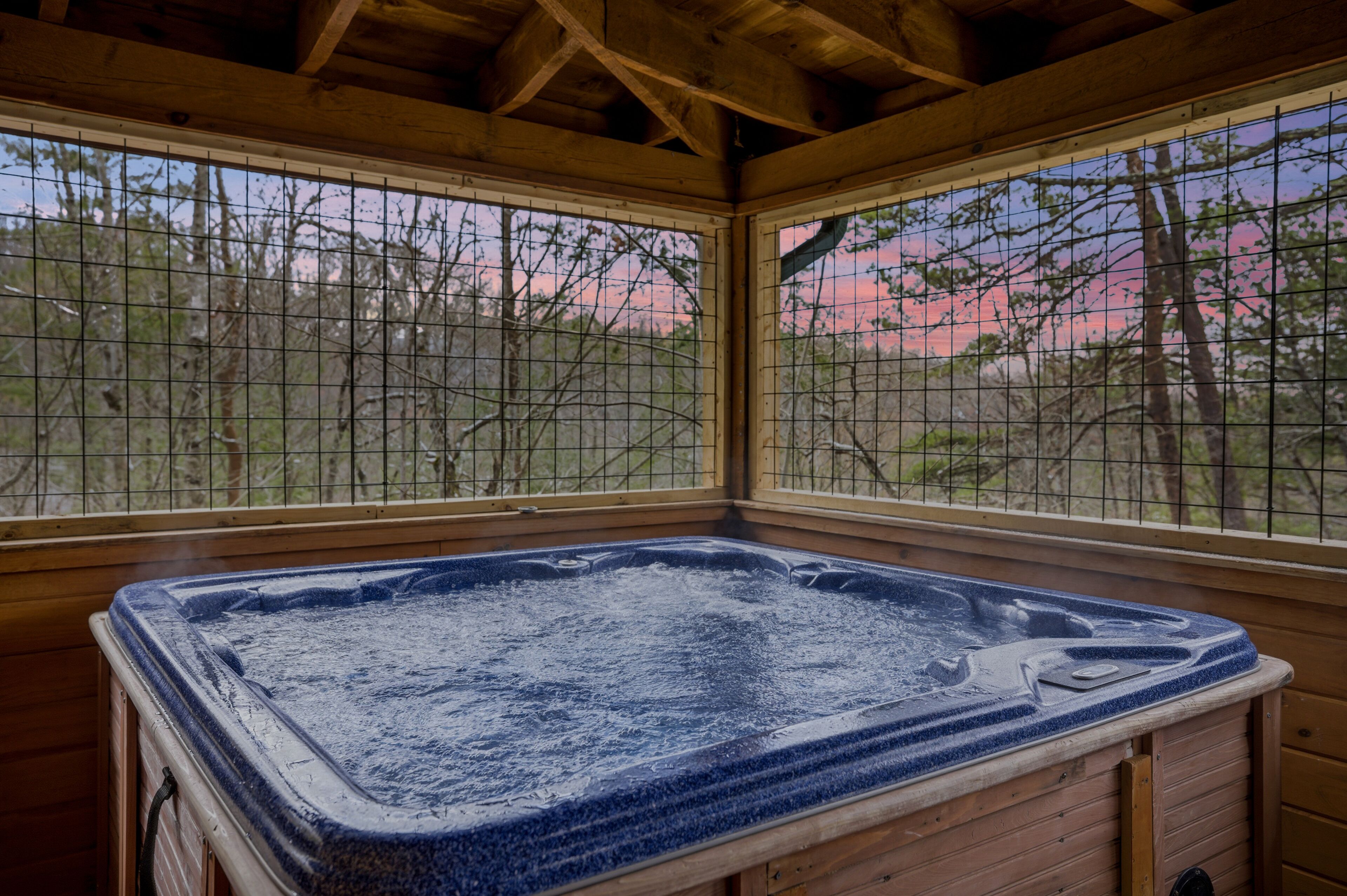 Ein Whirlpool in einer Holzhütte mit Blick auf Bäume und einem rosafarbenen Himmel.