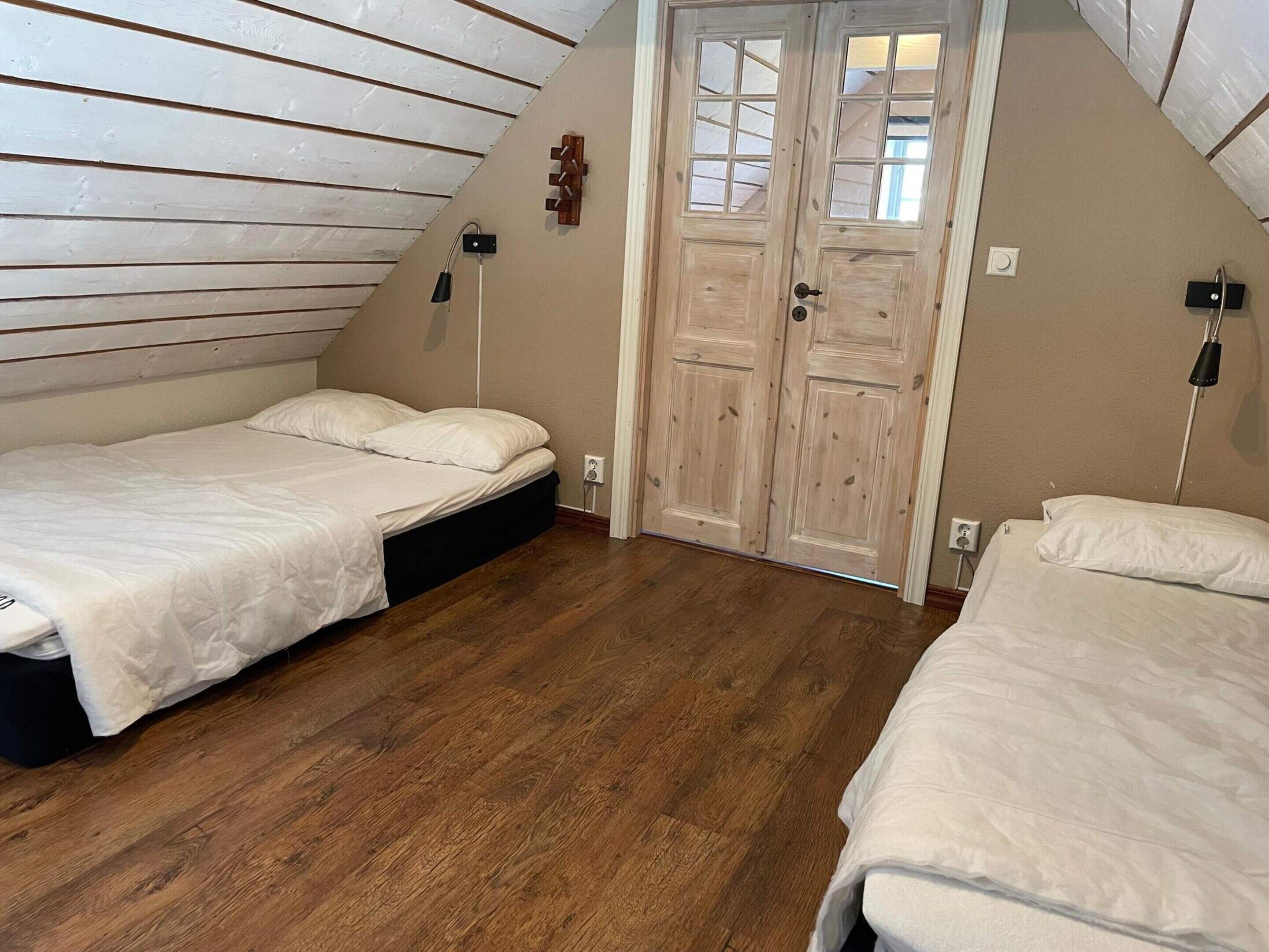 A small, cozy room with two beds, wooden flooring, a sloped ceiling, and a wooden door with glass panels.