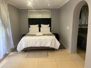 A bedroom with a bed, a nightstand, a window with curtains, and a door leading to another room. - Hazel Nest Ezulwini (Lobamba)