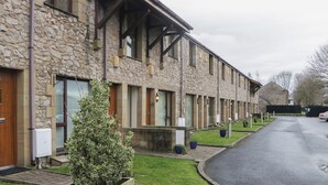 A row of stone buildings with wooden doors, a small tree in a planter, and a paved driveway. - 6 Swan House (Carnforth)