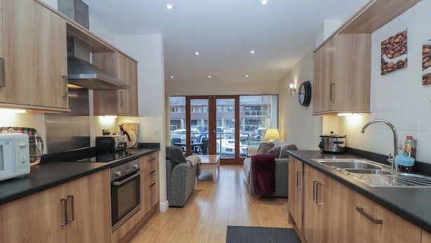 A modern kitchen with wooden cabinets, a built-in oven, and a large window overlooking a parking area. - 6 Swan House (Carnforth)