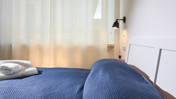 Ein Hotelzimmer mit Bett, Fenster mit durchsichtigen Vorhängen, Nachttischlampe und einer kleinen Pflanze auf dem Nachttisch.