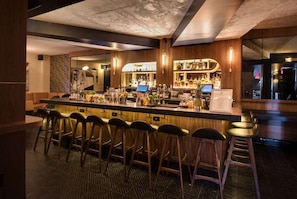 Una zona de bar con una barra alargada, varios taburetes y una gran variedad de botellas y vasos.