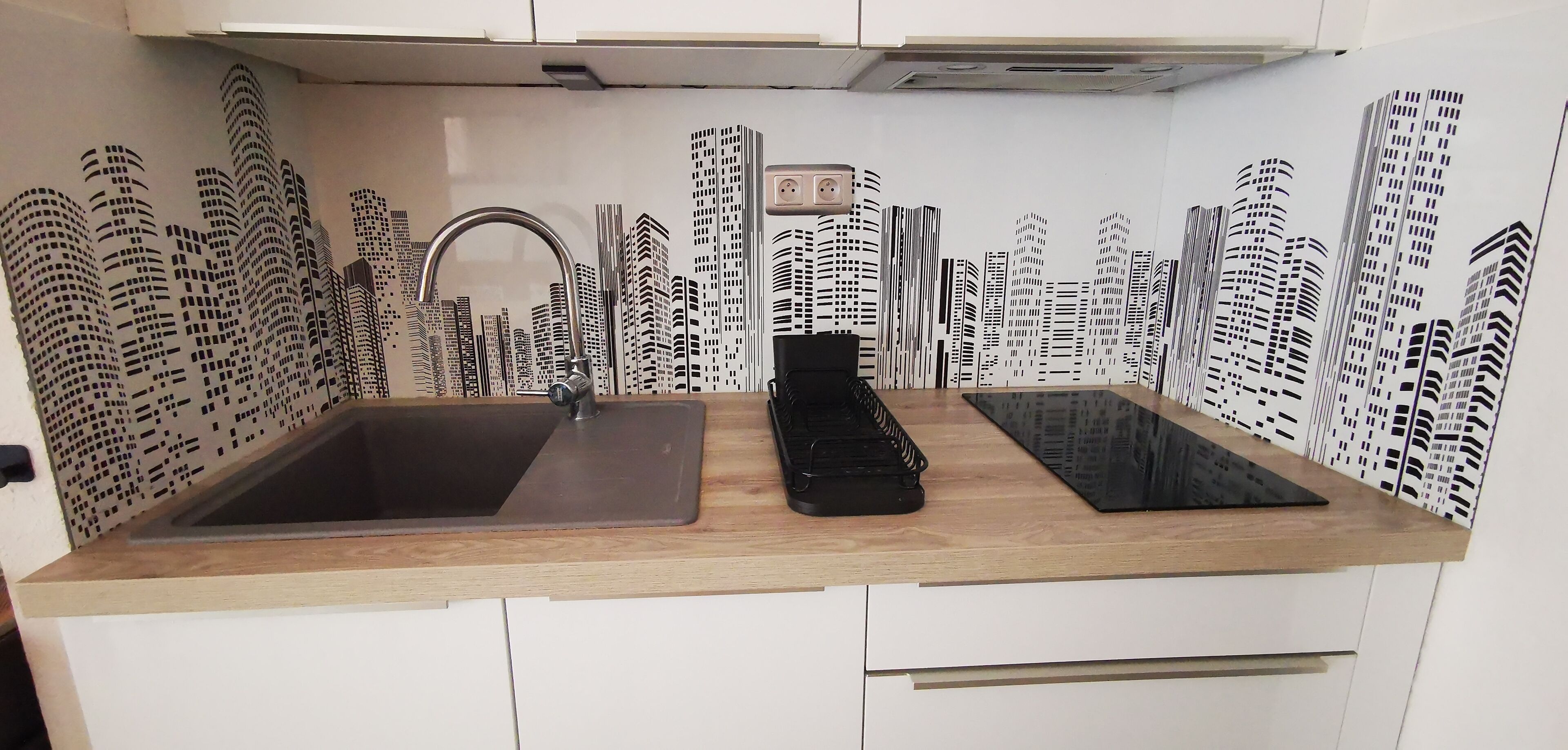 A kitchen with a double sink, a black electric kettle, and a backsplash with a cityscape design.