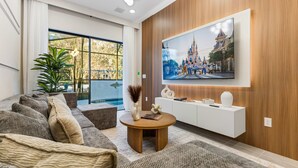 A modern living room with a large TV mounted on the wall, a comfortable sofa, a coffee table, and a view of a castle through the window. - 5BR 5BA Paradiso Grande - Walk to Seaworld 10507 (Orlando)