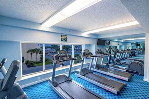 Une salle de sport équipée de plusieurs tapis de course, dotée d’une grande fenêtre et offrant une vue sur les palmiers à l’extérieur.