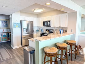 A modern kitchen with white cabinets, stainless steel appliances, a central island with stools, and a tiled backsplash. - Windy Hill Dunes 205 (North Myrtle Beach)