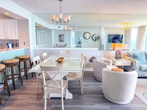 A modern kitchen with a dining area, a living room with a sofa and a TV, and a large mirror reflecting the space. - Windy Hill Dunes 205 (North Myrtle Beach)