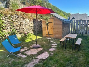 A wooden shed with a door, a red umbrella, a blue chair, and a small table with benches in a grassy yard. - The Patou's house (Cadéac)