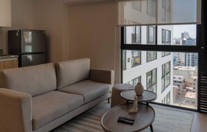 Ein modernes Wohnzimmer mit einer Couch, einem kleinen runden Tisch mit einer Vase und einer Fernbedienung, sowie einem großen Fenster mit Blick auf die Stadt.