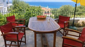Een houten tafel met zes rode stoelen, een mand met fruit en een gele parasol op een balkon met uitzicht op de kust.