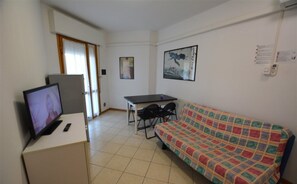 A small, sparsely furnished room with a bed, a desk with a computer, a television, and a door leading to another room. - Cozy Flat With Terrace and Pool Access (Lignano Sabbiadoro)