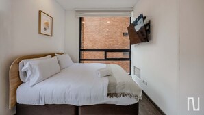 Quarto de hotel moderno com uma cama grande, uma TV de tela plana na parede e uma parede de tijolos visível pela janela.