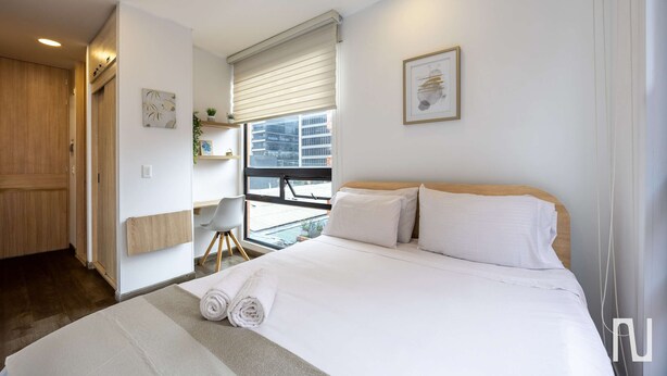 Quarto de hotel com uma cama grande, porta de madeira, janela com persianas e uma pequena mesa com uma cadeira.