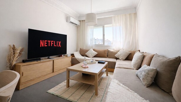 Moderni olohuone, jossa on litteänäyttöinen televisio, jonka ruudulla näkyy Netflixin logo, sohva, sohvapöytä ja suuret ikkunat, joista avautuu näkymä kaupunkiin.