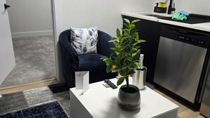 A modern living room with a white coffee table, a dark blue sofa, a potted plant, and a kitchen area with a dishwasher and a shelf above the counter. - Cozy 1 bedroom Suite, Kitchenette & self check in (Edmonton)