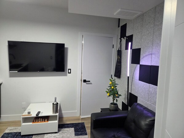 Un salon moderne avec un téléviseur à écran plat fixé au mur, une petite table basse sur laquelle est posé un jeu d’échecs, une plante en pot et un canapé de couleur sombre.