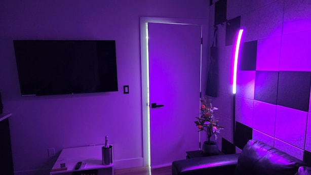 A modern living room with a flat-screen TV mounted on the wall, a white coffee table with a bottle, a black leather sofa, and a door with purple lighting. - Cozy 1 bedroom Suite, Kitchenette & self check in (Edmonton)