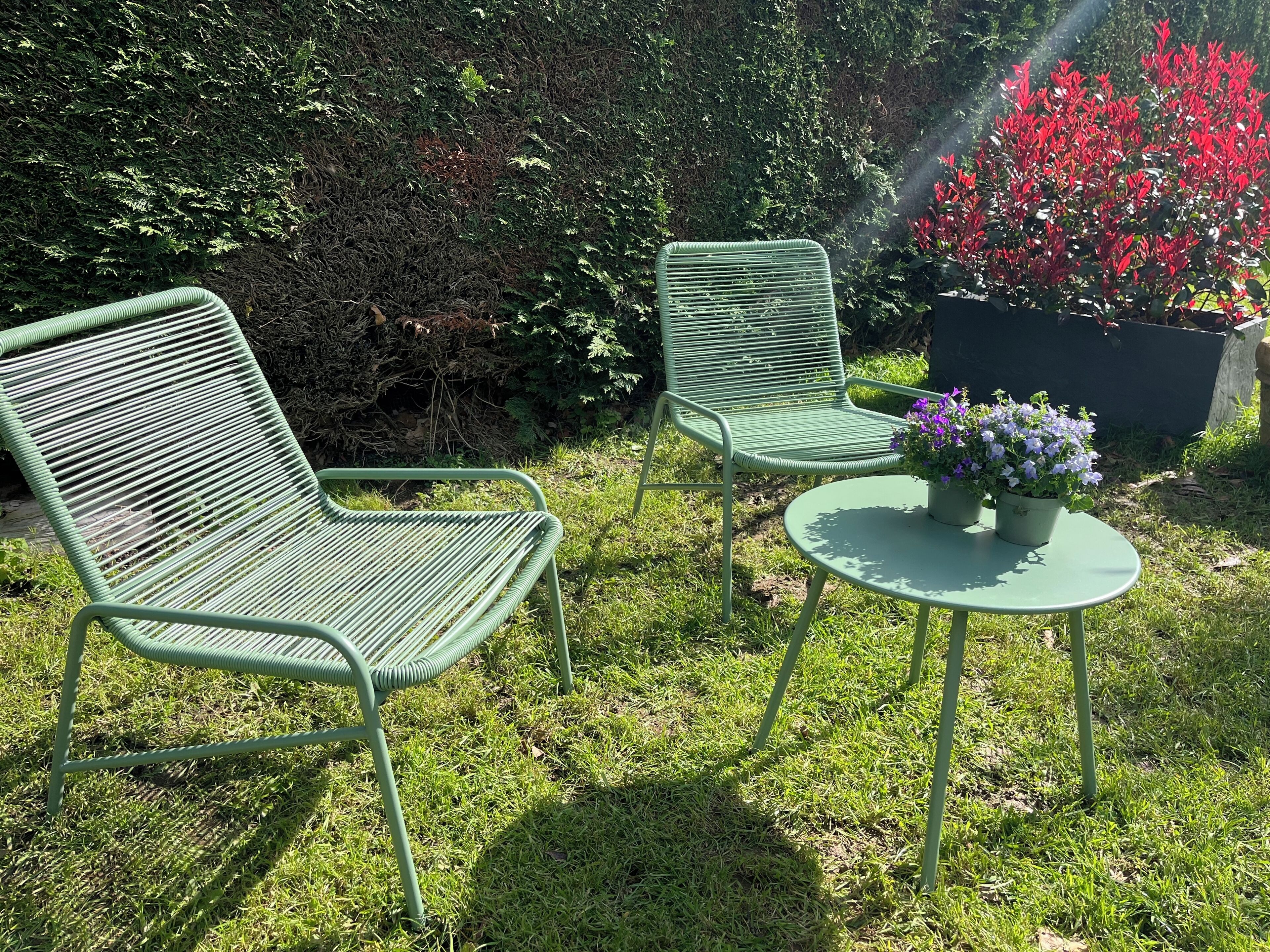 Un juego de silla y mesa metálicos verdes en un jardín con césped y una siega al fondo.