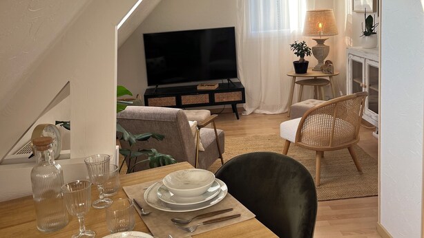 A dining area with a wooden table set for four, a television in the background, and a cozy seating arrangement with a sofa and chairs. - Le Petit Cosy - King Size Bed, Quiet & Parking (Vaucelles)