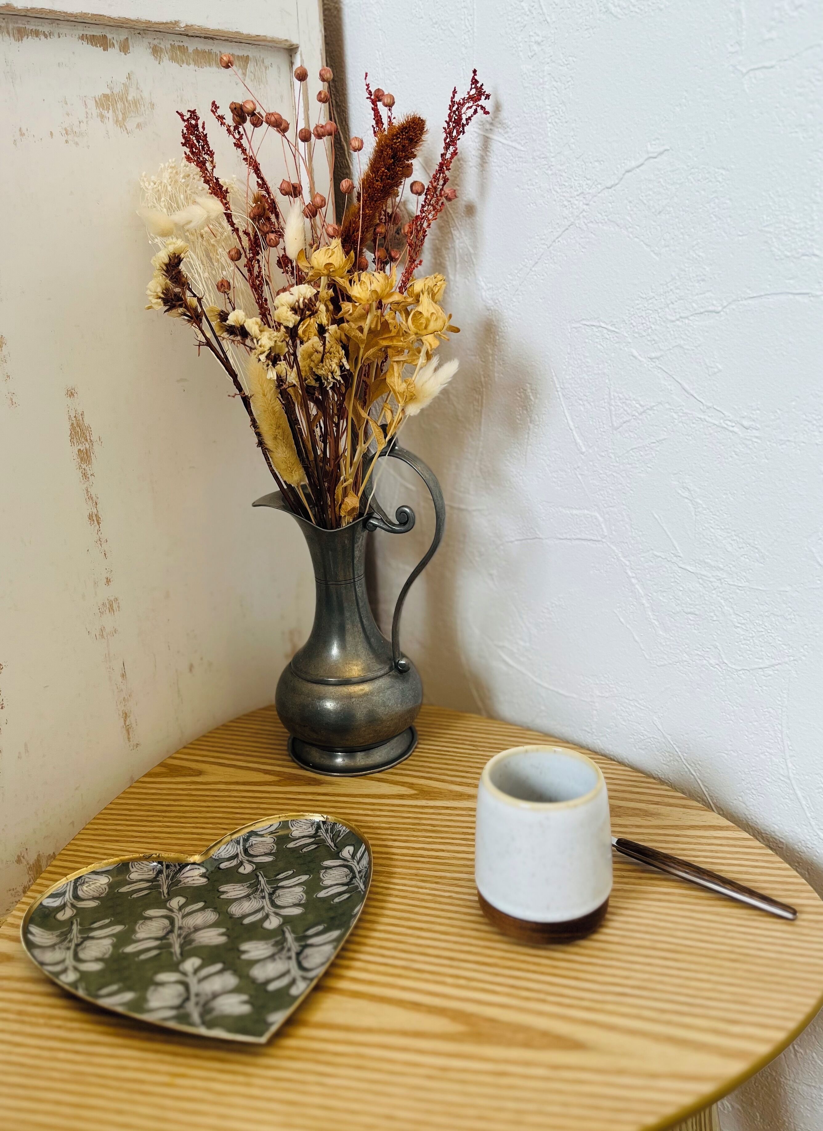 Una mesa en esquina con un florero de metal con flores secas, un plato de cerámica y una taza blanca.
